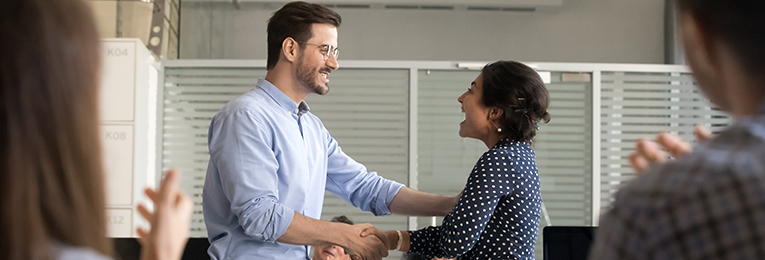 A person expressing appreciation to their coworker by shaking hands in an office space, while people in the foreground applaud.