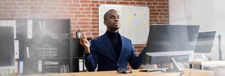 A person in a suit is seen relaxing with their eyes closed and hands above their waist at their office desk.