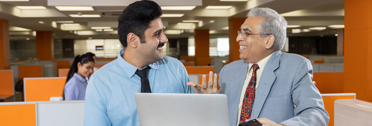 A senior individual in a gray suit and a younger person, both smiling, engaged in discussion at a work table.