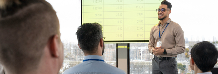 A person stands by a screen displaying statistics, addressing their team members during an office meeting.
