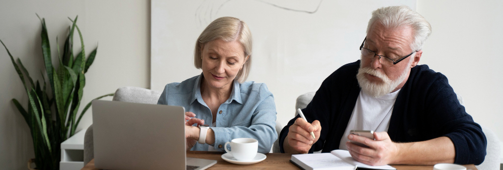 A senior-aged couple is seen calculating at a desk with a laptop, coffee cup, and notes spread out.