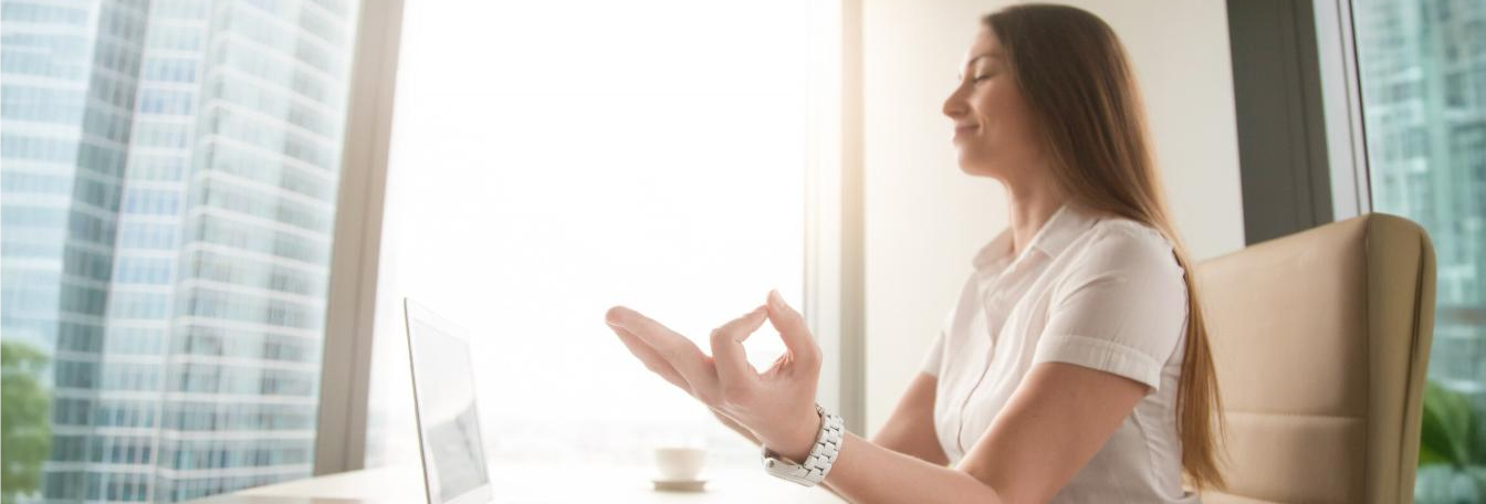 A woman is seen meditating in front of her desk and laptop, her hands facing upward in a tranquil and relaxed position.