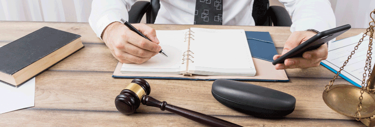 A person's lower half is seen taking notes in a notebook while referring to their phone, with a gavel and weighing scale placed on the table.