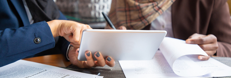 Midsection view of a woman referencing a hand-held device while explaining something to a colleague beside her, who is holding several documents.